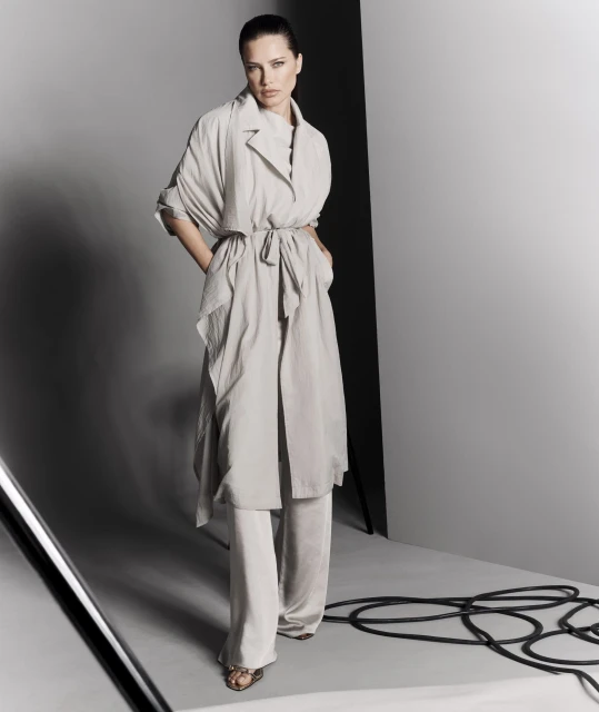 Full-length shot of a woman in a light-colored belted coat and pants posing in a studio with gray walls and black cables on the floor.