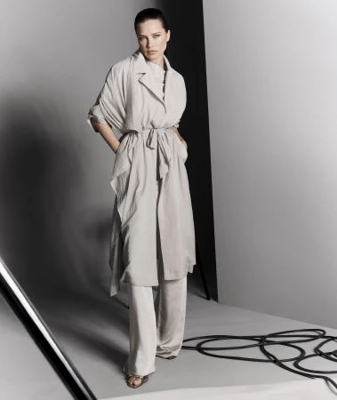 Full-length shot of a woman in a light-colored belted coat and pants posing in a studio with gray walls and black cables on the floor.