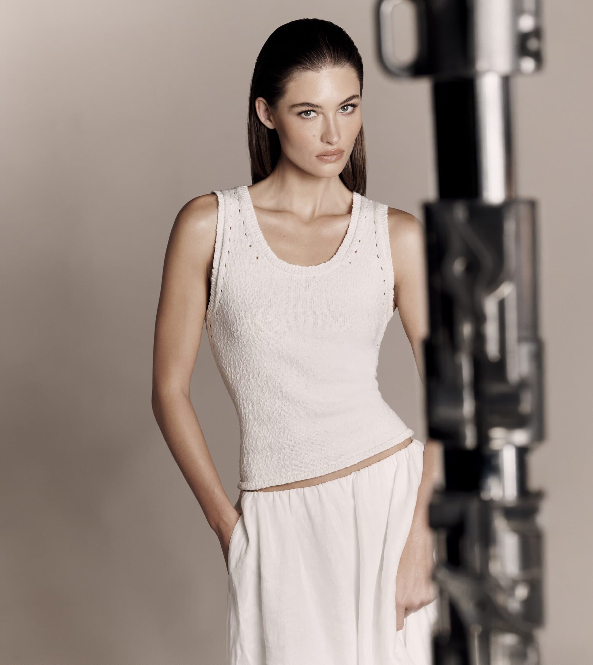 A slender brunette woman poses in a studio, wearing a textured white tank top and wide-leg pants, standing next to a blurred light stand.