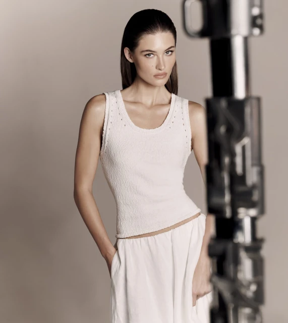 A slender brunette woman poses in a studio, wearing a textured white tank top and wide-leg pants, standing next to a blurred light stand.