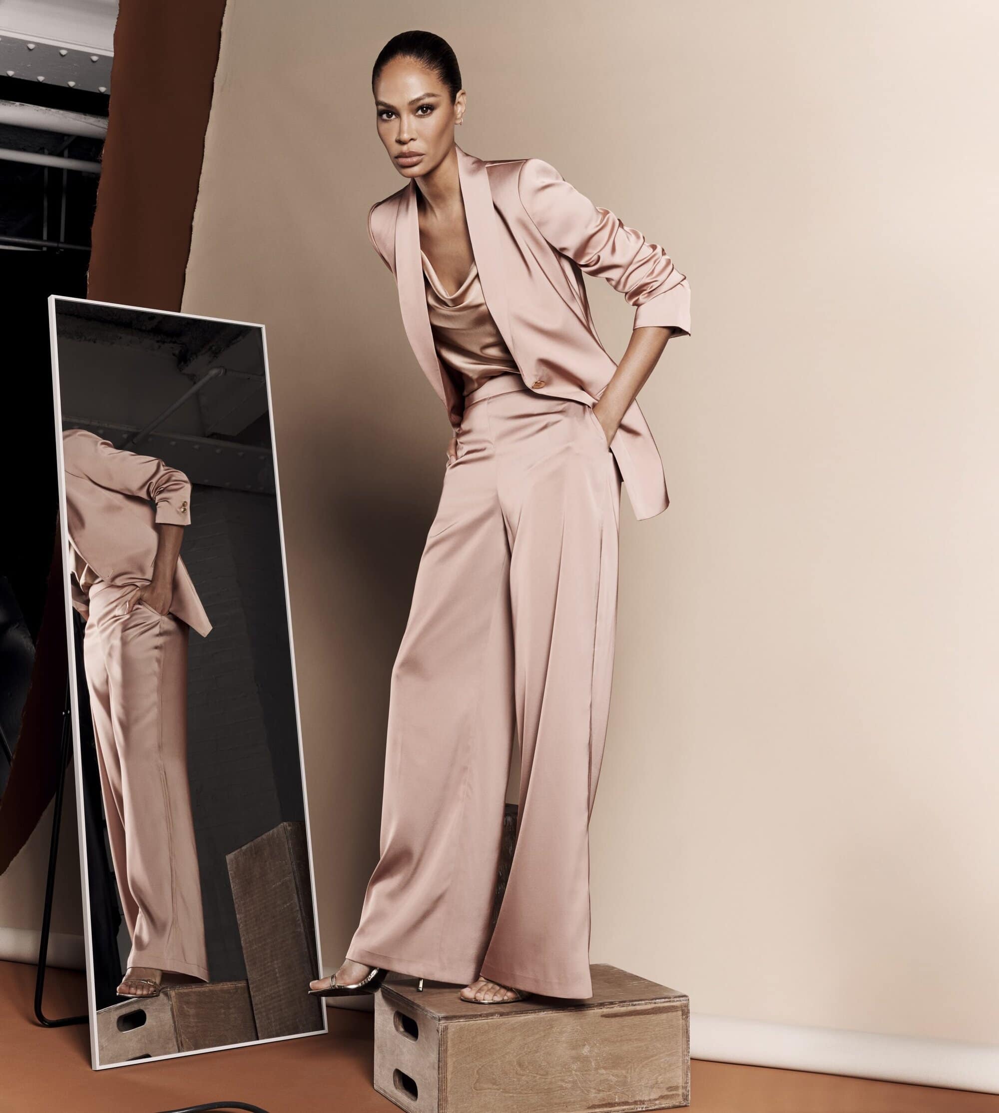 Elegant woman in rose-colored satin suit poses on wood block in neutral studio with mirror reflection. Fashion, style, and beauty concept.