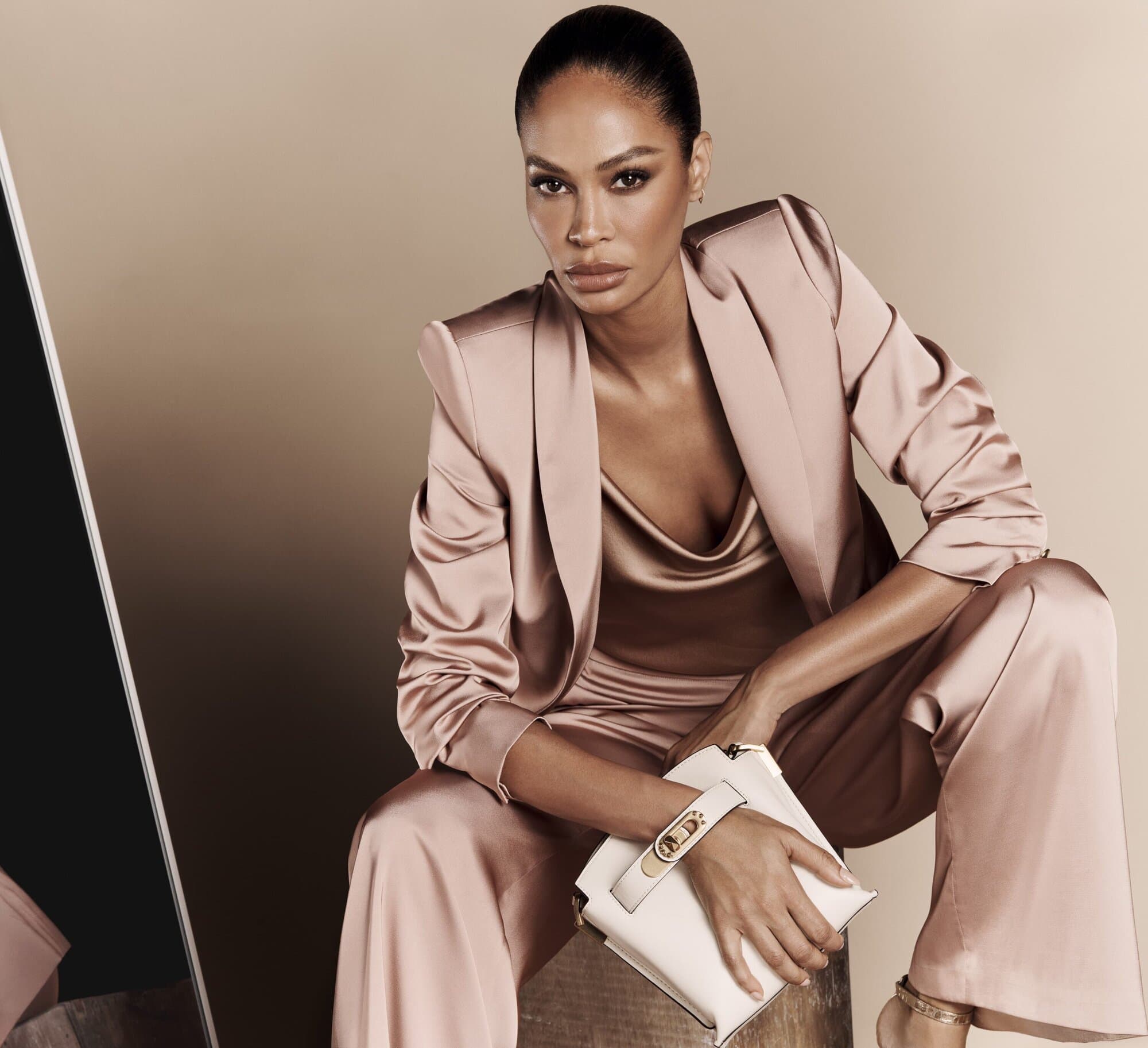 Elegant adult female in a satin suit with a clutch, seated indoors against a tan background, accented by a mirror on the left.