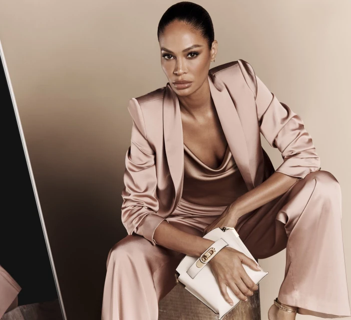 Elegant adult female in a satin suit with a clutch, seated indoors against a tan background, accented by a mirror on the left.