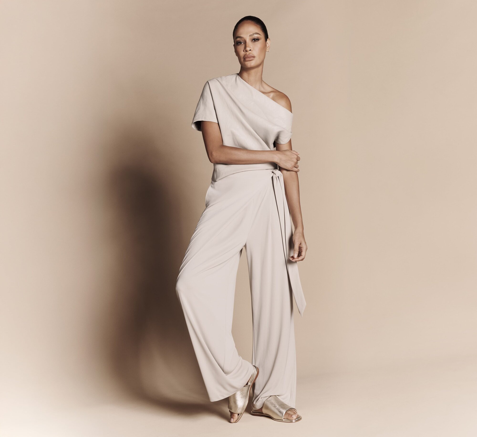 Full-length portrait of an adult female fashion model in a light neutral-tone jumpsuit and sandals posing in front of a neutral background.