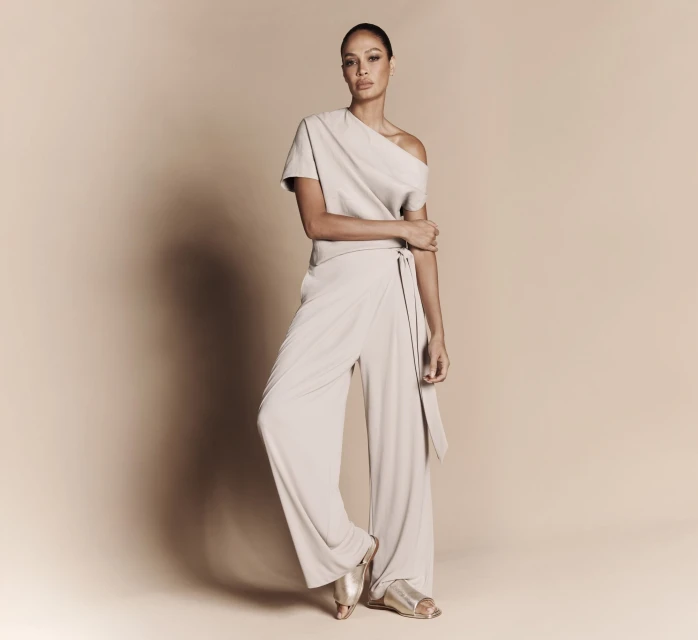 Full-length portrait of an adult female fashion model in a light neutral-tone jumpsuit and sandals posing in front of a neutral background.