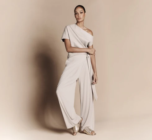 Full-length portrait of an adult female fashion model in a light neutral-tone jumpsuit and sandals posing in front of a neutral background.