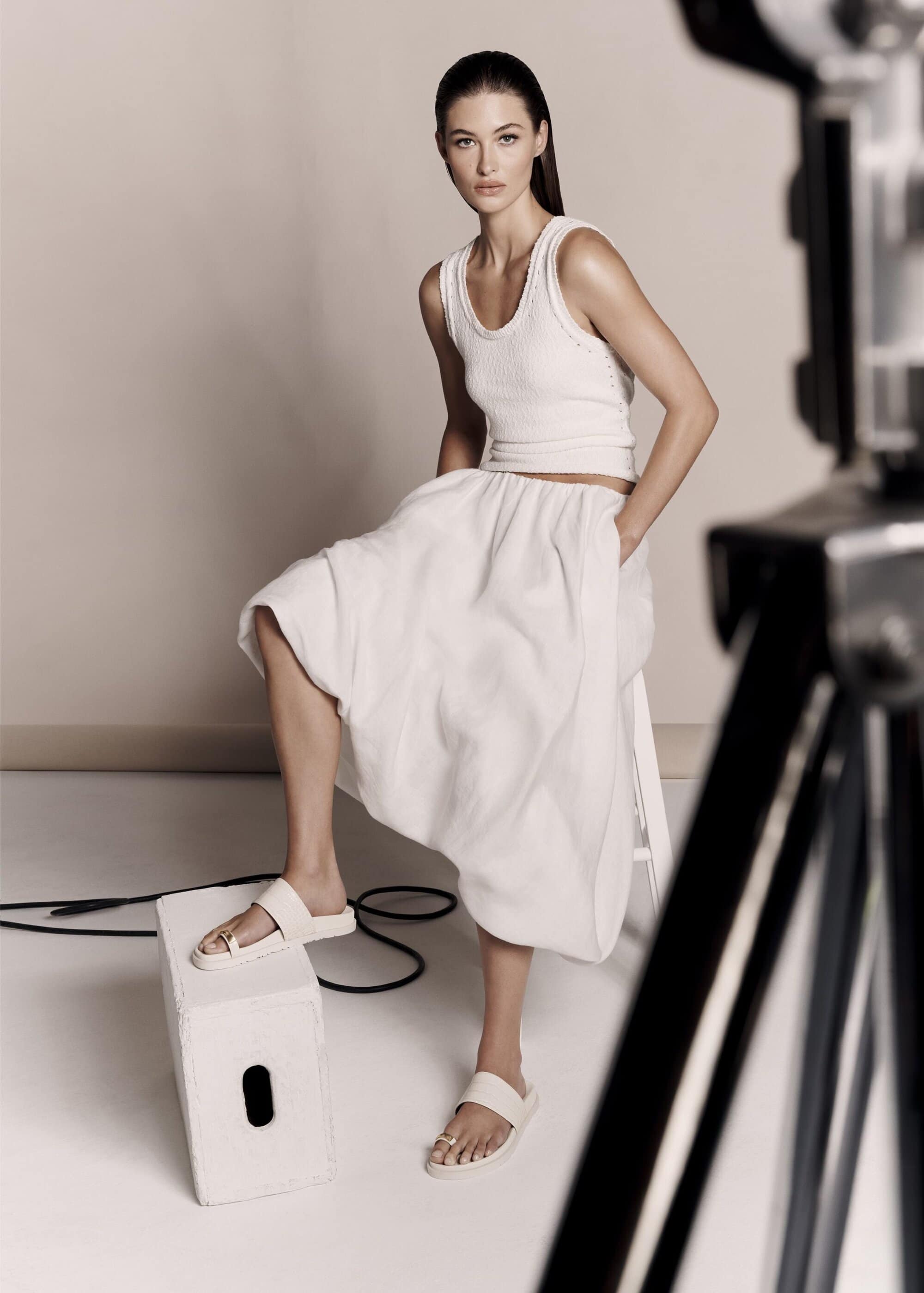 A fair-skinned adult woman poses in white separates and slide sandals in a studio with a softbox light visible in the frame.