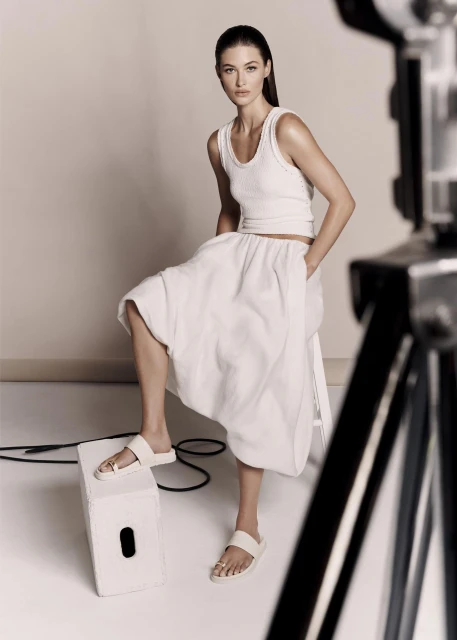 A fair-skinned adult woman poses in white separates and slide sandals in a studio with a softbox light visible in the frame.