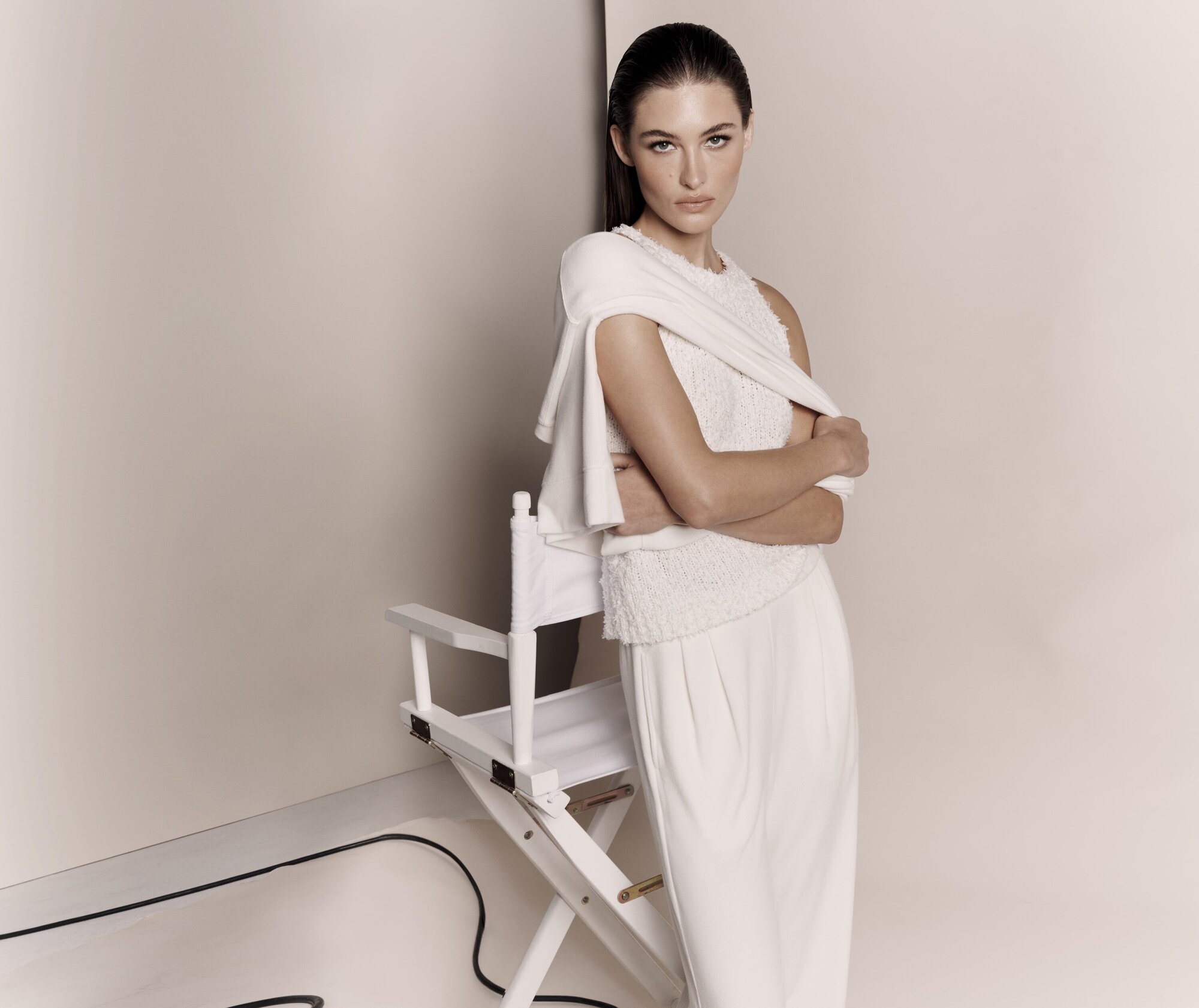 Elegant woman in an all-white outfit poses in front of a white background, standing next to a white director's chair in a fashion studio.