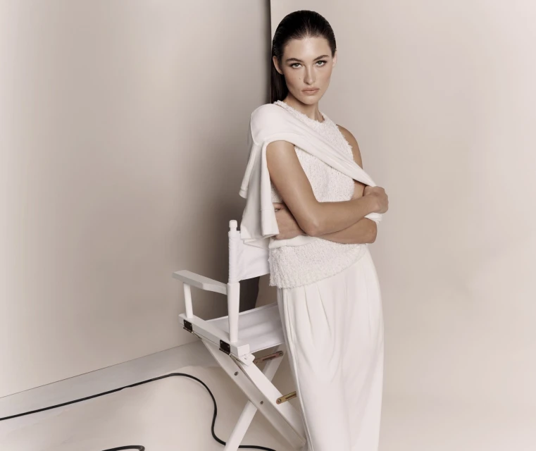 Elegant woman in an all-white outfit poses in front of a white background, standing next to a white director's chair in a fashion studio.
