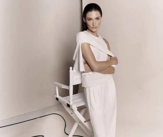 Elegant woman in an all-white outfit poses in front of a white background, standing next to a white director's chair in a fashion studio.