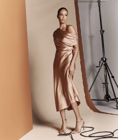Elegant adult female poses in a studio wearing a light brown off-shoulder dress and strappy heels, illuminated by studio lighting.