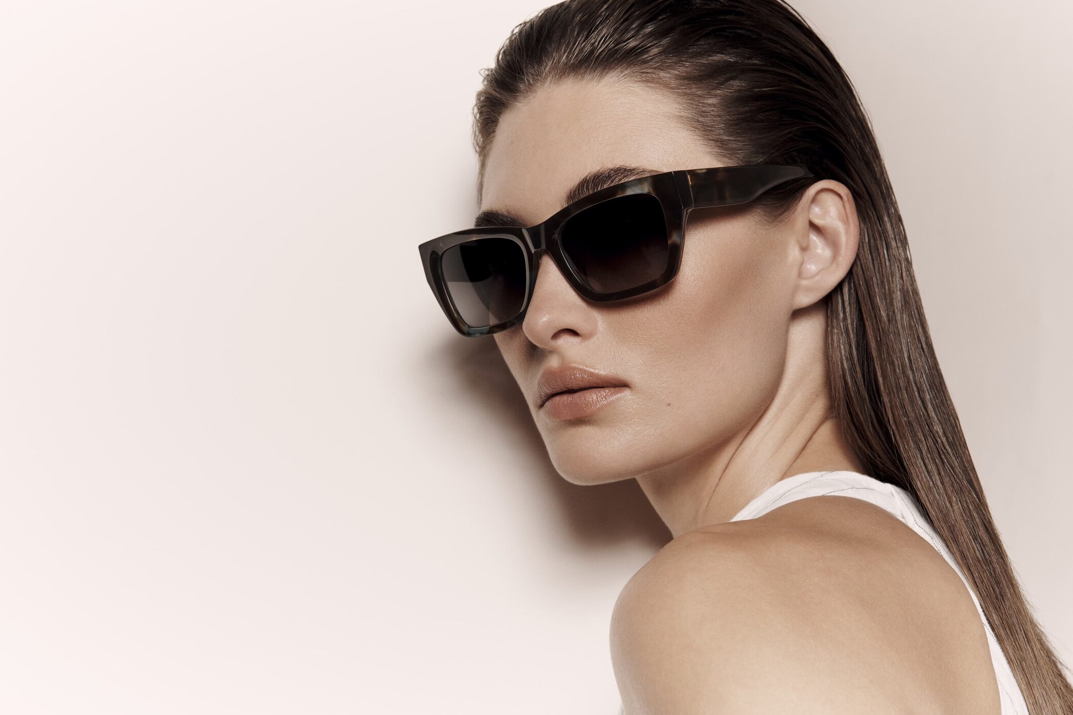 Close-up portrait of a woman wearing sunglasses, with slicked-back hair, a neutral expression, against a neutral, solid background.