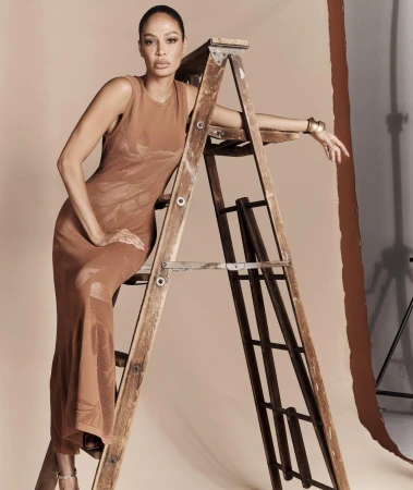 Model leaning against a wooden ladder wearing a sleeveless brown knit dress in a studio setting.