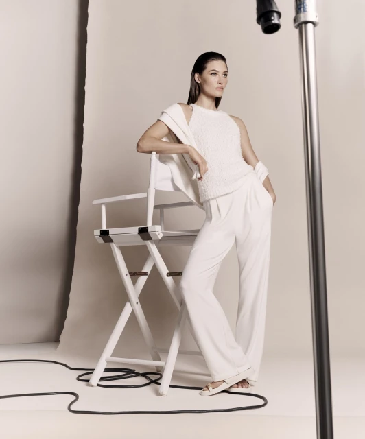 Woman in white textured top and pants poses in a photo studio, leaning against a director's chair with a light stand in the background.