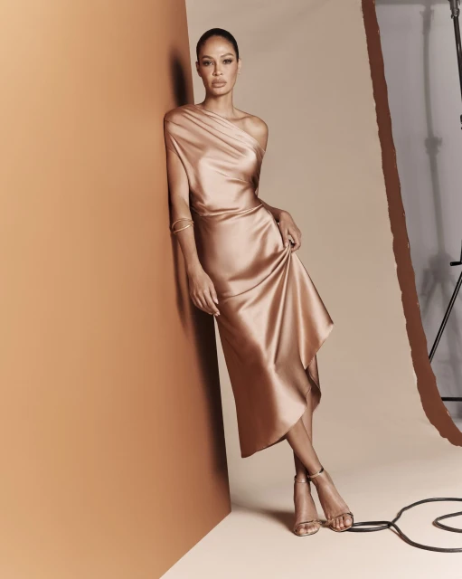 Elegant adult female poses in a champagne-colored, one-shoulder dress with high heels against a neutral backdrop in a fashion studio.