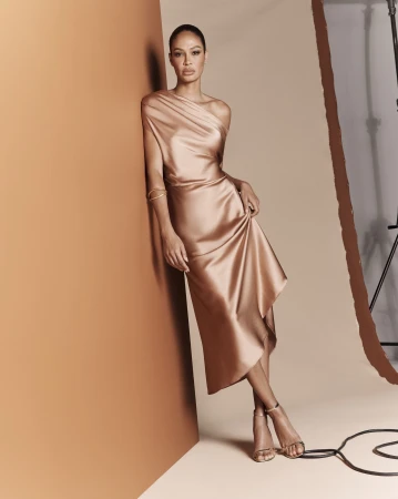 Elegant adult female poses in a champagne-colored, one-shoulder dress with high heels against a neutral backdrop in a fashion studio.