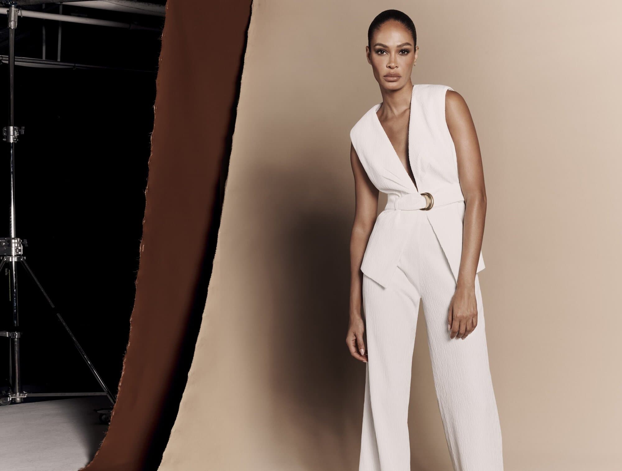 Model Joan Smalls wearing a white sleeveless belted jumpsuit against a studio seamless background