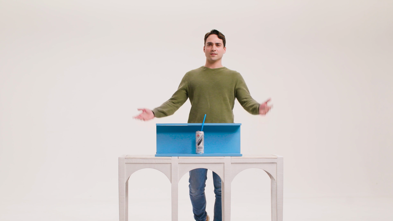Man in green shirt stands behind light blue table with drink can and blue straw in white studio, spreading arms out.