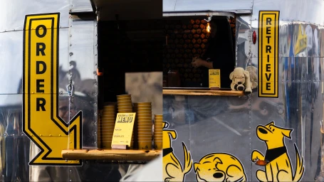 Silver food truck exterior with yellow order and retrieve signs, stacks of yellow cups, and illustrated dog graphics for Hertz campaign.