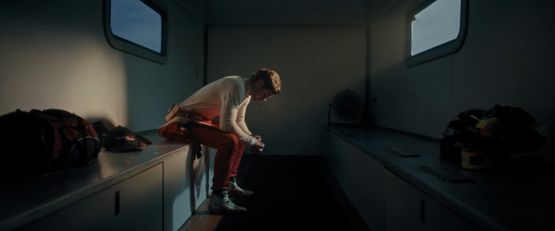 Man in racing suit sits pensively in a dimly lit room near helmet and duffel bag, illuminated by light from a window.