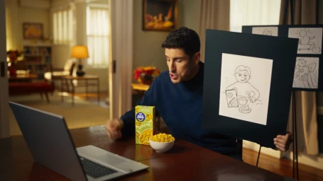 Man holding a black presentation board with a drawing while seated at a wooden table with a laptop and a Kraft Mac and Cheese box.