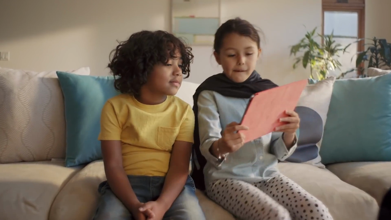 Two children sitting on a living room sofa looking at a red tablet in a warm interior setting