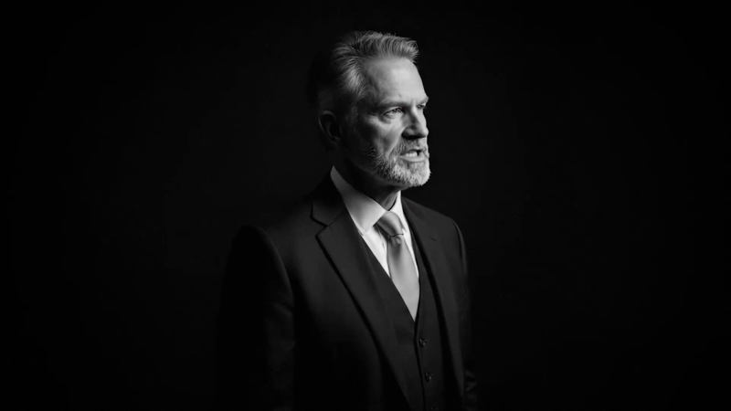 Monochrome headshot of a mature man in a suit and tie looking off camera against a dark background in a studio setting.