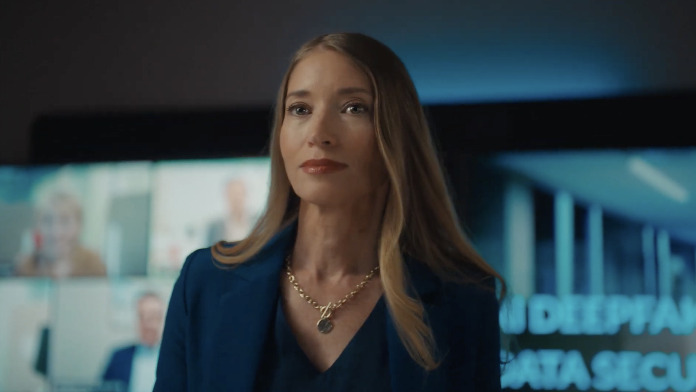 Portrait of a blonde adult woman wearing a navy blue blazer and gold necklace, looking off-camera in an office environment.