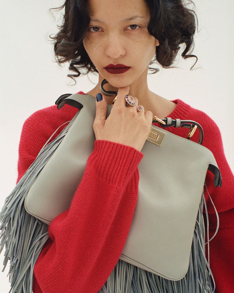 Adult woman in a red sweater holding a grey leather fringe purse and wearing statement rings against a white backdrop.