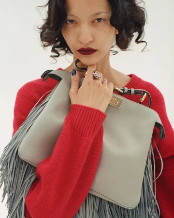 Adult woman in a red sweater holding a grey leather fringe purse and wearing statement rings against a white backdrop.