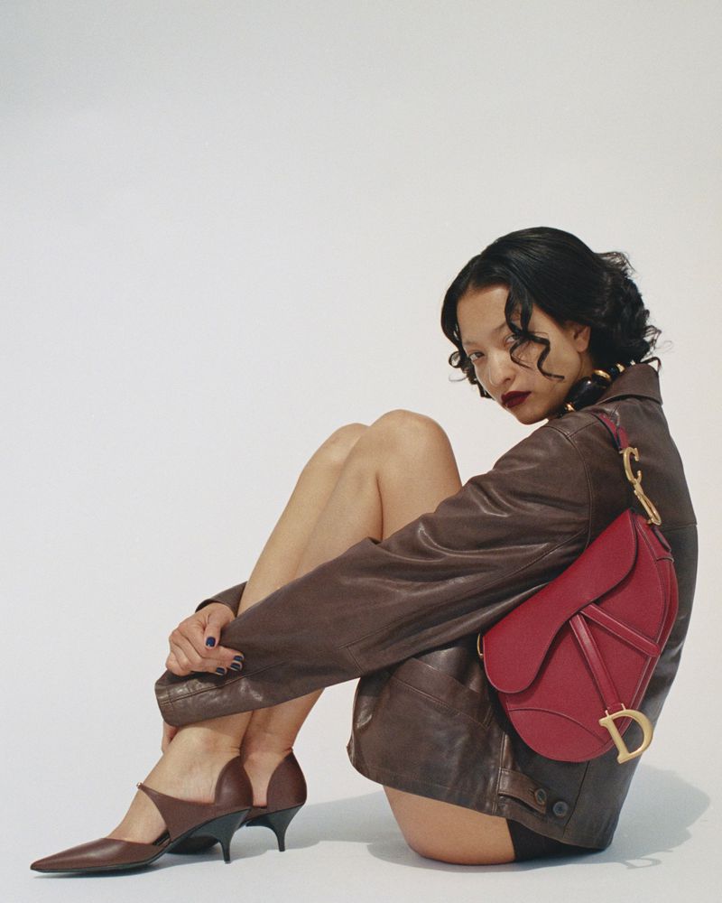 Elegant adult woman poses in a brown jacket, dark shoes, and a red handbag, seated in a photo studio with neutral white background.