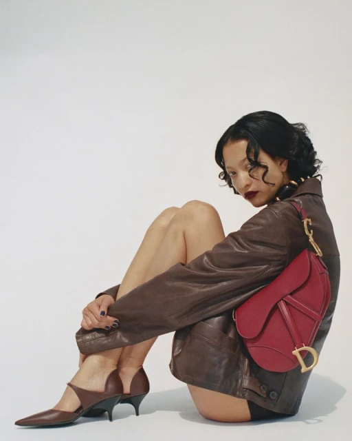 Elegant adult woman poses in a brown jacket, dark shoes, and a red handbag, seated in a photo studio with neutral white background.
