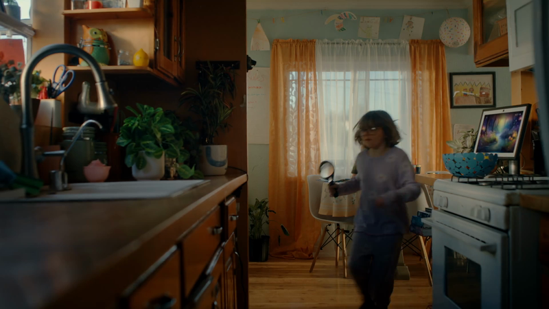 A child explores the kitchen with a magnifying glass, standing near the oven and countertop. The room features orange curtains and playful artwork.