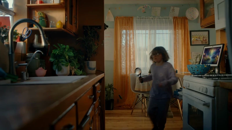 A child explores the kitchen with a magnifying glass, standing near the oven and countertop. The room features orange curtains and playful artwork.