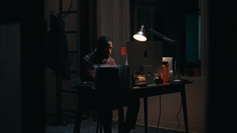 A man works at a desktop computer in a dimly lit room with a desk lamp, computer, and water bottle visible. He is wearing headphones.