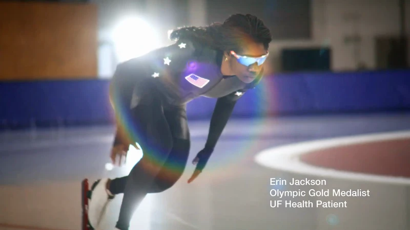 Female speed skater Erin Jackson in action on an indoor ice rink wearing a black suit with an American flag, stars, and blue mirrored glasses.