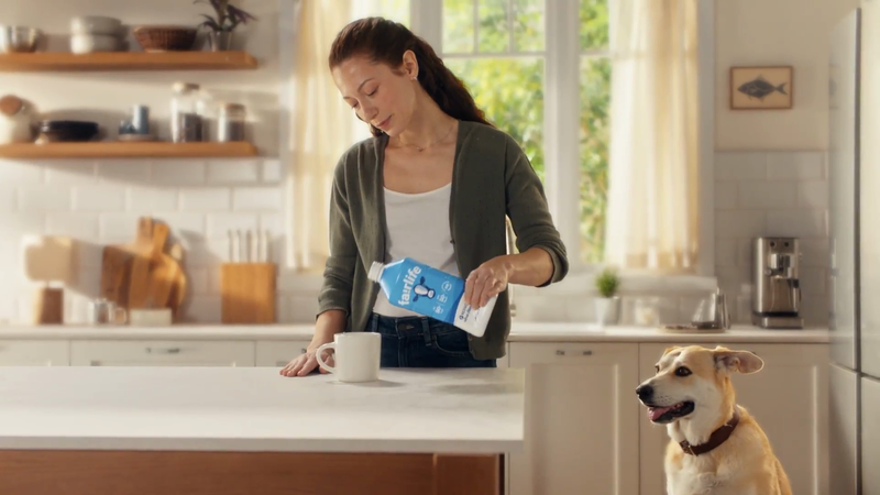Woman pours plant-based milk into a mug in a bright kitchen as a dog watches. Interior scene with white cabinets and natural light.