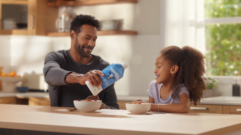 Father pouring milk into daughter's cereal bowl at the kitchen table; a sweet and healthy breakfast scene in a bright, modern home.