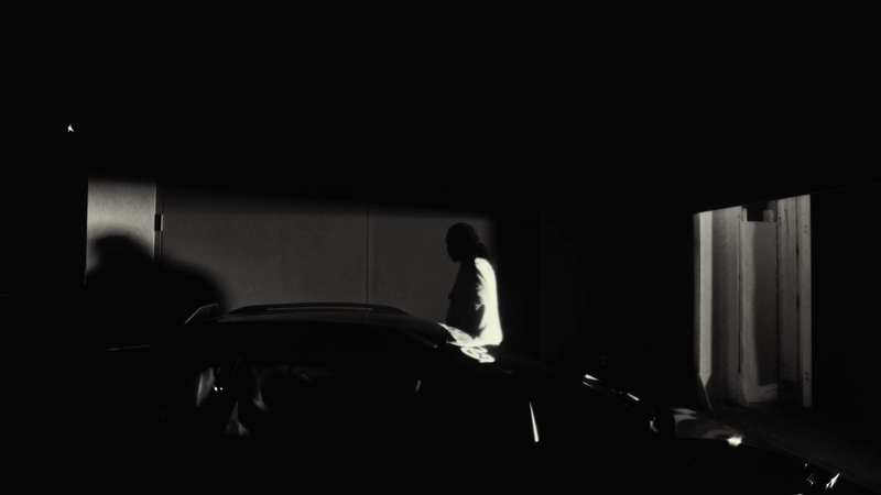Person in white jacket stands near dark car in a dimly lit, indoor parking garage with dark shadows and black and white tones.