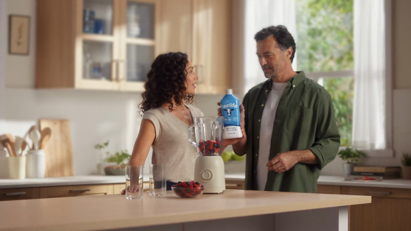 Couple in a bright kitchen preparing a smoothie with fresh berries, using a blender and a carton of plant-based milk.