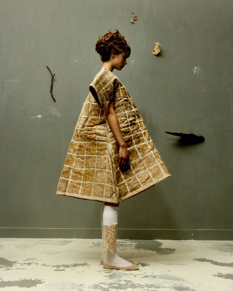 A person stands in a gray room wearing a quilted tan and white dress with matching leg coverings in a studio setting.