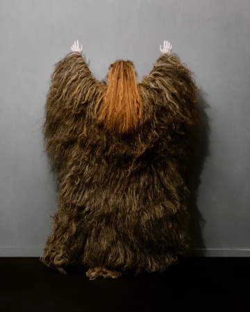 An abstract shot of a person with orange hair covered with a shaggy brown fur-like cape or coat standing against a grey wall.