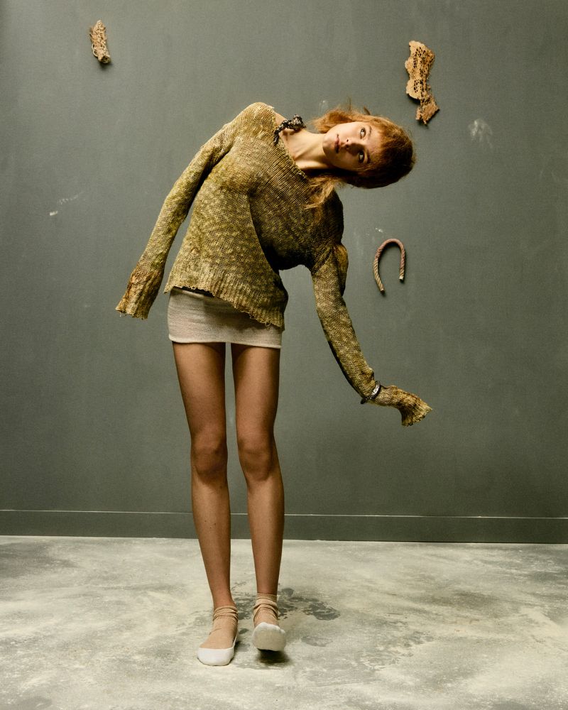 A redhead woman in a tan-and-brown sweater and white mini skirt poses indoors against a grey wall with decorative horseshoe and wood pieces.