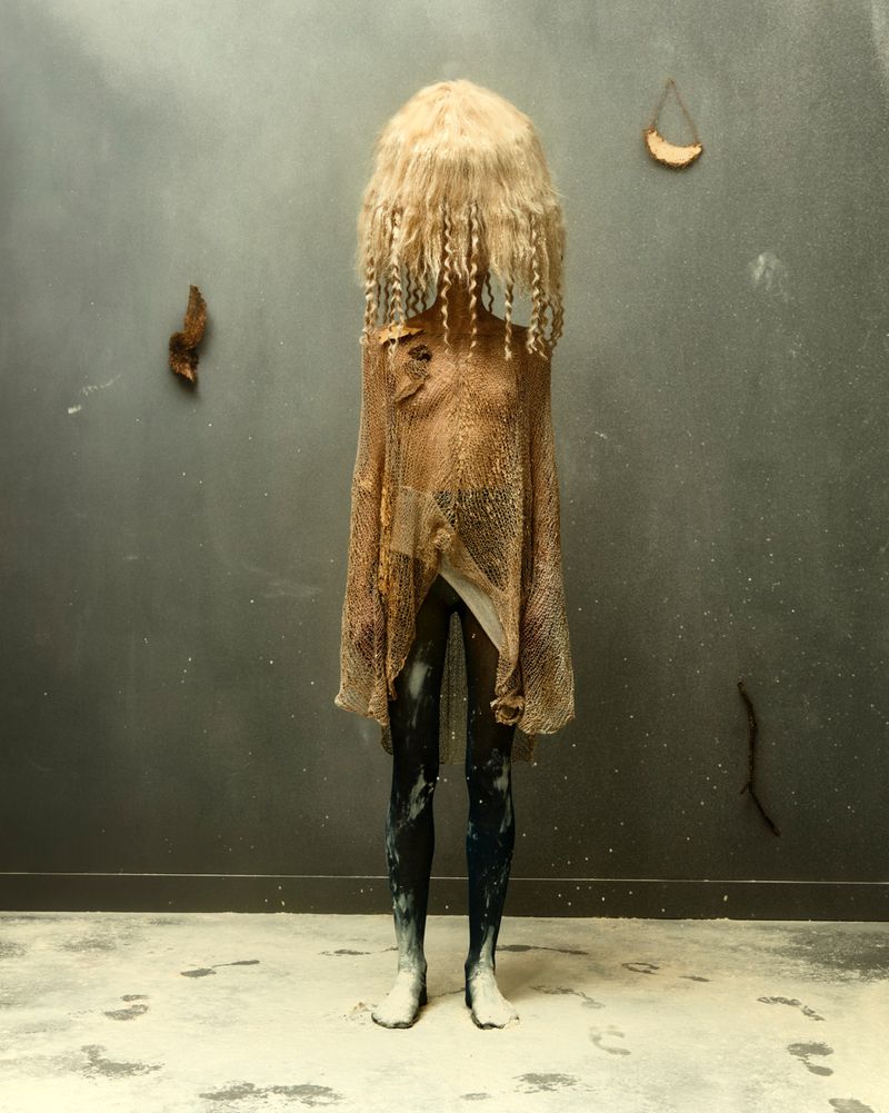 Full-length artistic photo features a person with a blonde wig and draped fabric in front of a gray wall, creating an eerie and unique look.