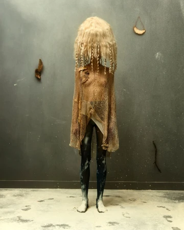 Full-length artistic photo features a person with a blonde wig and draped fabric in front of a gray wall, creating an eerie and unique look.
