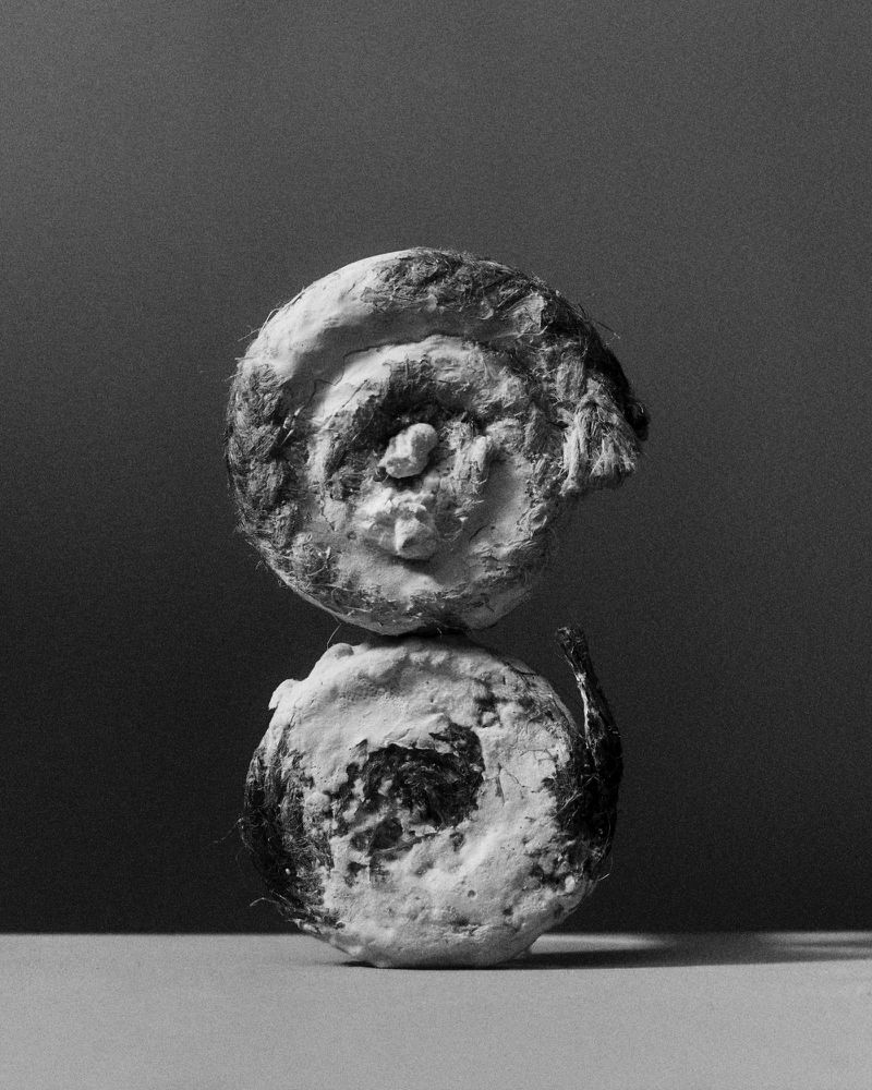 Monochrome still life features two round objects stacked vertically, with spiral patterns and rough textures against a dark background.