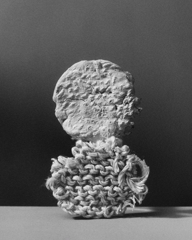 Monochrome studio still life of a plaster bust fragment balanced on a textured, knitted object, set against a smooth grey backdrop.