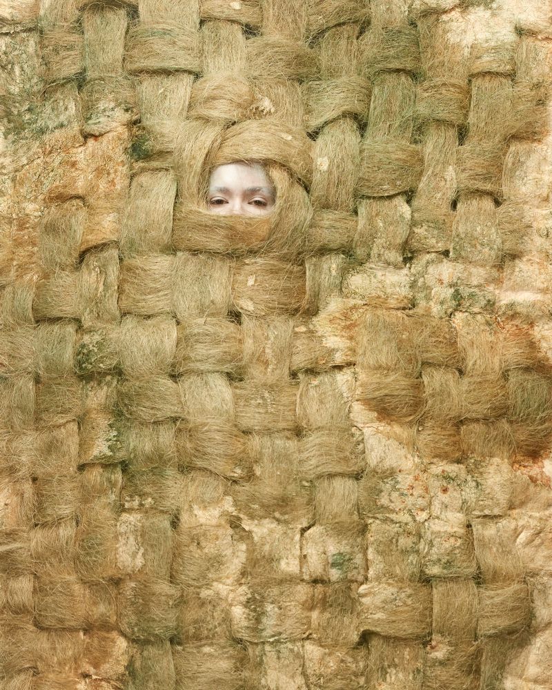 Abstract portrait of a person's face peering through woven material, creating a textured and earthy composition, in a studio setting.