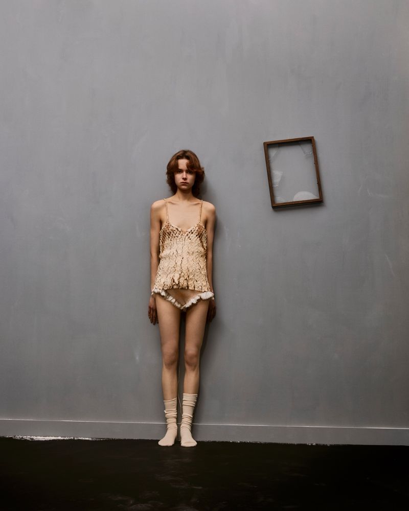 Young woman in lacy lingerie stands against a gray wall, near a floating frame, in a minimalist fashion shot with soft light.